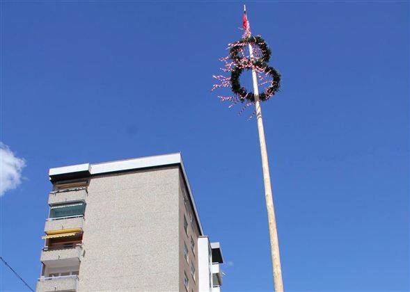 Vysoký stožár s vlajkou, věncovými a barevnými světly pod jasně modrou oblohou. V pozadí je vidět obytná budova.