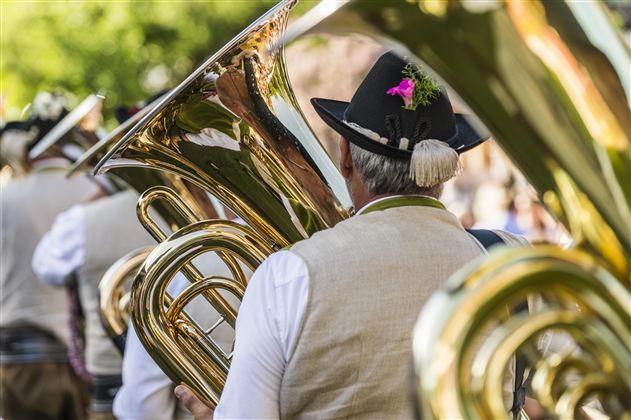 A group of musicians with tubas is wearing traditional clothing. In the background, trees and a festive atmosphere can be seen.