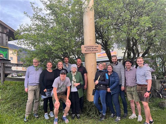 Skupina lidí stojí před kmenem stromu se značkou. Usmívají se a pózují v zeleném prostředí.