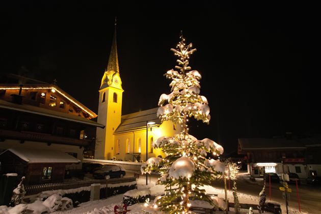 Ein festlich geschmückter Weihnachtsbaum steht im Schnee vor einer beleuchteten Kirche. Die Szenerie ist nachts und strahlt eine gemütliche Stimmung aus.
