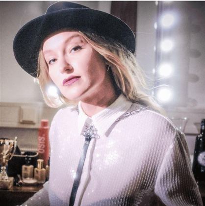 Eine stilvolle Person mit einem Hut und einem eleganten Outfit steht vor einem beleuchteten Spiegel. Die Atmosphäre wirkt glamourös und modern.