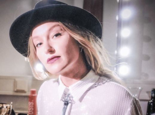 Eine Frau mit blonden Haaren trägt einen schwarzen Hut und steht vor einem beleuchteten Spiegel. Sie hat ein selbstbewusstes Lächeln und trägt ein glitzerndes Oberteil.