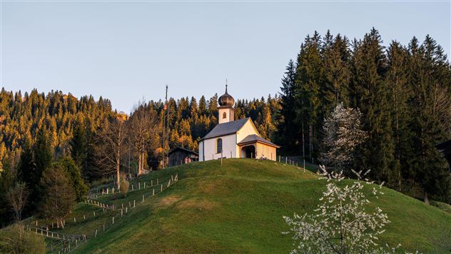 Eine kleine Kirche auf einem Hügel, umgeben von Bäumen. Die Landschaft ist grün und die Atmosphäre ist ruhig.