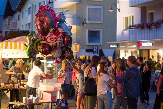 Eine belebte Stadtstraße mit einer Menschenmenge und Ständen. Ein bunter Ballon mit einer Figur zieht die Aufmerksamkeit auf sich.