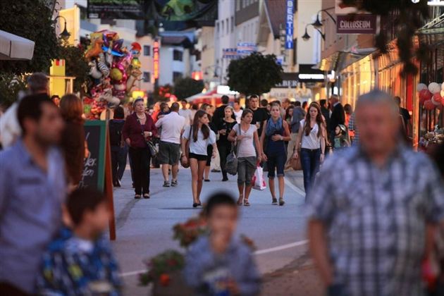 Eine belebte Fußgängerzone mit vielen Menschen, die einkaufen und spazieren gehen. Bunte Schilder und Stände sorgen für eine lebhafte Atmosphäre.