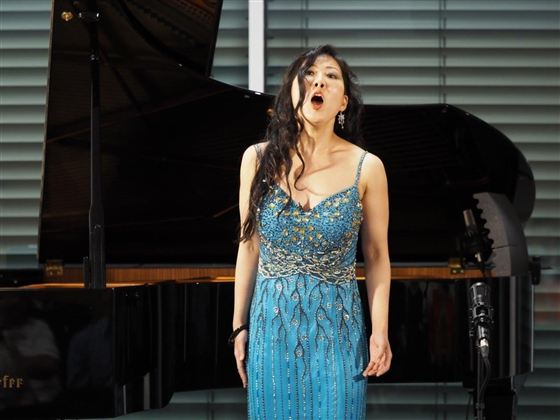 Eine Sängerin in einem blauen, glitzernden Kleid steht vor einem Klavier und singt leidenschaftlich. Im Hintergrund sind Fenster mit horizontalen Jalousien sichtbar.