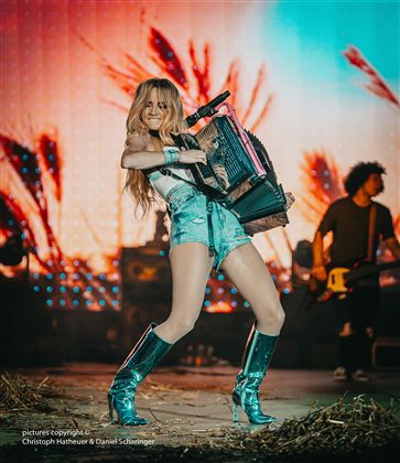 Eine junge Frau mit langen Haaren spielt Akkordeon auf einer Bühne. Im Hintergrund sind Lichter und Silhouetten von Musikern zu sehen.