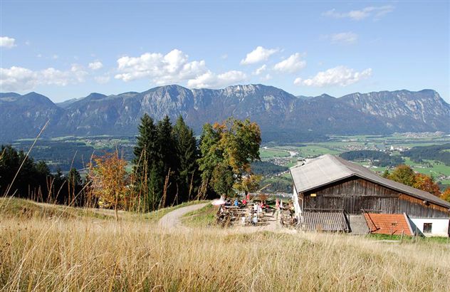 Eine malerische Landschaft mit Bergen im Hintergrund und einer Wiese im Vordergrund. Ein rustikales Gebäude ist sichtbar, umgeben von Bäumen und einem klaren Himmel.