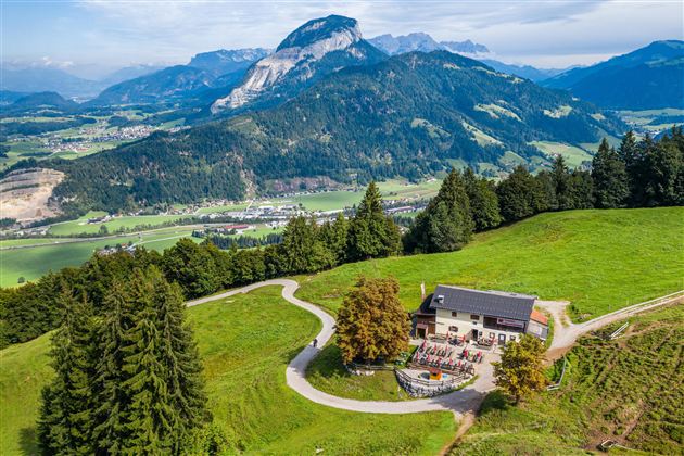 Eine malerische Landschaft mit einem gemütlichen Haus, umgeben von Bäumen und Wiesen. Im Hintergrund erheben sich majestätische Berge unter einem blauen Himmel.