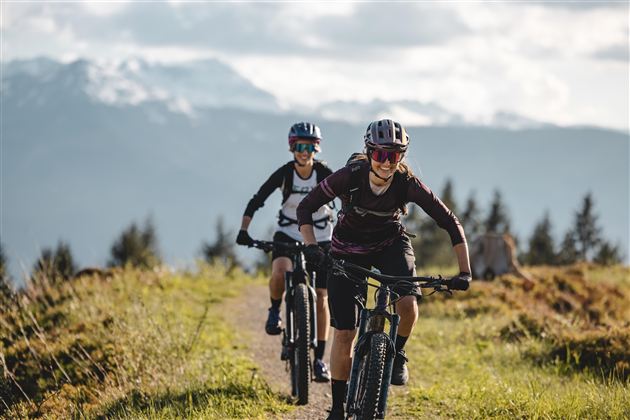 Zwei Radfahrer fahren auf einem Trampelpfad in den Bergen. Im Hintergrund sind schneebedeckte Gipfel und eine bewaldete Landschaft zu sehen.