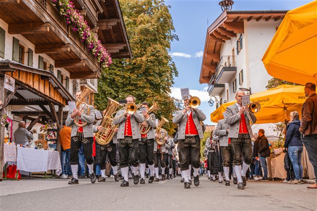 A music band in traditional costumes marches through a festive street. In the background, stands and colorful sun umbrellas can be seen.