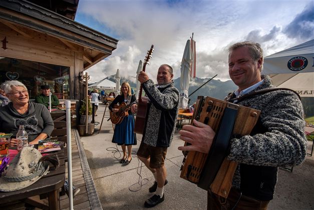 Eine Gruppe von Musikern spielt traditionelle Musik vor einer Berghütte. Im Hintergrund sind tanzende Menschen und eine malerische Berglandschaft zu sehen.