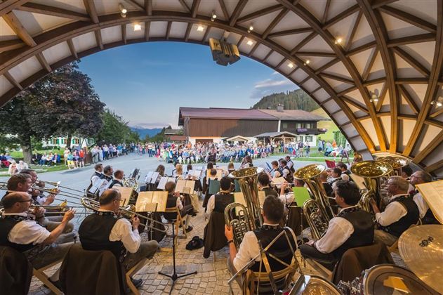 Eine Blaskapelle spielt vor einer großen Menschenmenge unter einem gewölbten Dach. Im Hintergrund sind Berge und ein klarer Himmel sichtbar.