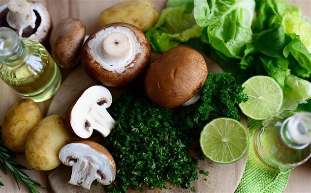 Výběr čerstvých ingrediencí na dřevěné desce, včetně hub, brambor, salátu a limetek. Patří sem také olivový olej a čerstvé bylinky.