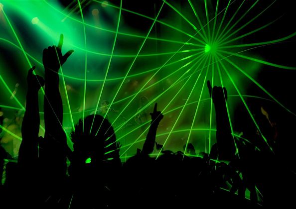 Een menigte met opgeheven handen, omgeven door groene lichtstralen. De sfeer is levendig en feestelijk.