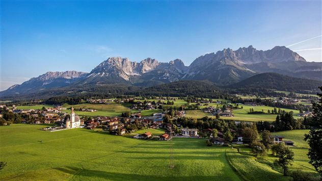 A picturesque landscape with green meadows and a small village. In the background, majestic mountains rise under a clear sky.