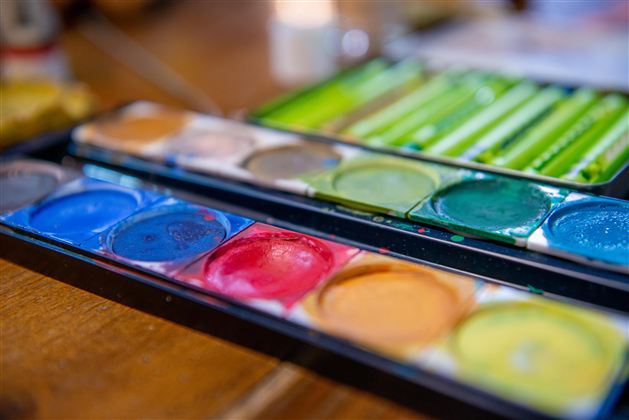 Eine Palette mit Wasserfarben in verschiedenen Farben liegt auf einem Tisch. Im Hintergrund sind unscharfe Gegenstände zu sehen.