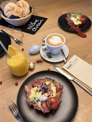 Ein köstliches Frühstück mit Rinderbraten, pochiertem Ei und Kirschtomaten auf einem schwarzen Teller. Im Hintergrund sind eine Tasse Kaffee und ein Glas Orangensaft zu sehen.