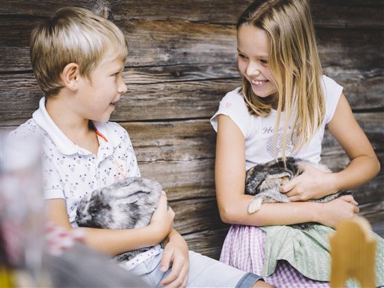 Zwei Kinder sitzen nebeneinander und lächeln sich an. Sie halten jeweils ein Kaninchen auf dem Schoß.
