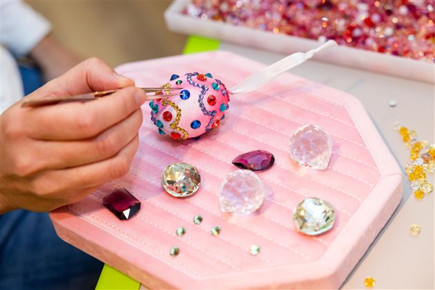 A hand decorates a colorful egg with rhinestones. Next to it are various sparkling stones on a pink surface.
