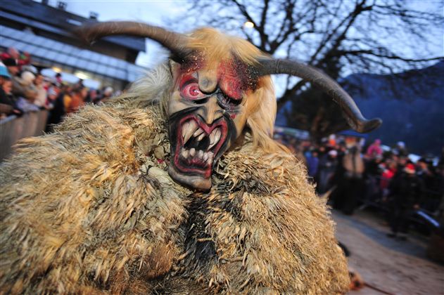 Eine Person trägt ein gruseliges Kostüm mit einer Maske und einem Fellumhang. Im Hintergrund sieht man eine Menschenmenge und Bäume.