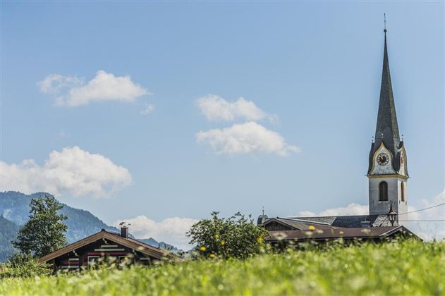 Eine malerische Landschaft mit einer Kirche und einem traditionellem Gebäude im Vordergrund. Der Himmel ist blau mit einigen Wolken und die Berge sind im Hintergrund sichtbar.