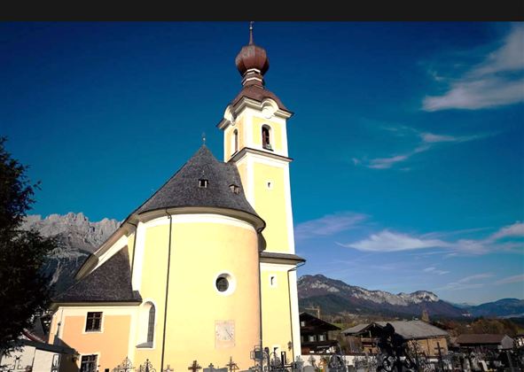 A beautiful church with a yellow facade and a distinctive tower. In the background, mountains and a blue sky can be seen.