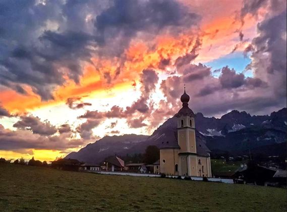 A picturesque church against a spectacular evening backdrop. The sky is filled with vibrant colors and clouds, while the mountains are visible in the background.