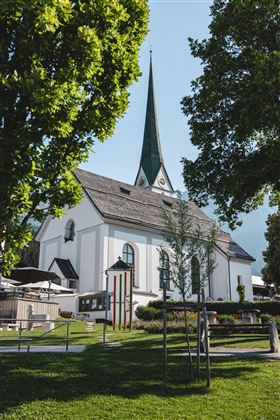 Eine schöne Kirche mit einem hohen, spitzen Turm. Umgeben von Bäumen und einer grünen Wiese.