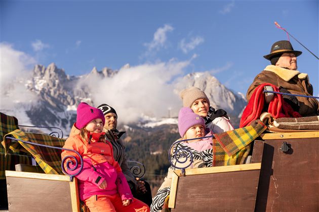 Eine fröhliche Familie auf einer Pferdeschlittenfahrt in den Bergen. Im Hintergrund sind schneebedeckte Gipfel und ein klarer Himmel zu sehen.