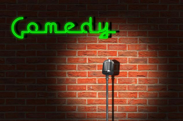 Ein Mikrofon steht auf einer Bühne vor einer Backsteinwand. Darüber leuchtet das Wort "Comedy" in grüner Neonfarbe.