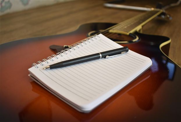 Ein Notizblock mit einem Stift liegt auf einer Akustikgitarre. Der Hintergrund ist unscharf und zeigt eine gemütliche Umgebung.