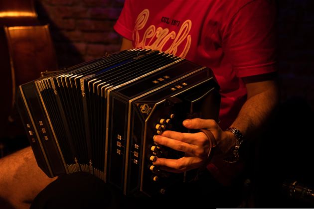 Ein Musiker spielt ein Bandoneon in einem gedämpften, warmen Licht. Seine Hand bewegt sich über das Instrument, während im Hintergrund eine Mauer sichtbar ist.