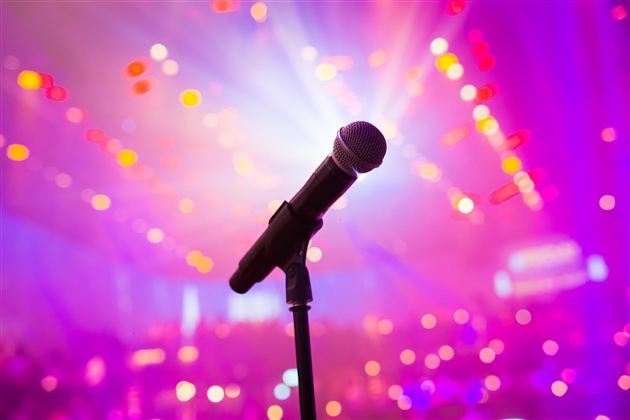 Ein Mikrofon steht im Fokus vor einem bunten, verschwommenen Lichtspiel. Es vermittelt eine festliche Stimmung und den Eindruck eines live Events.