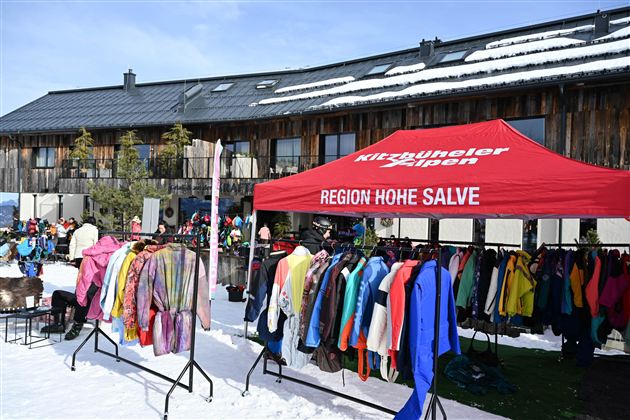 Ein Verkaufsstand mit bunten Ski- und Wintersportoutfits steht im Schnee. Im Hintergrund ist ein Gebäude mit Balkonen zu sehen.