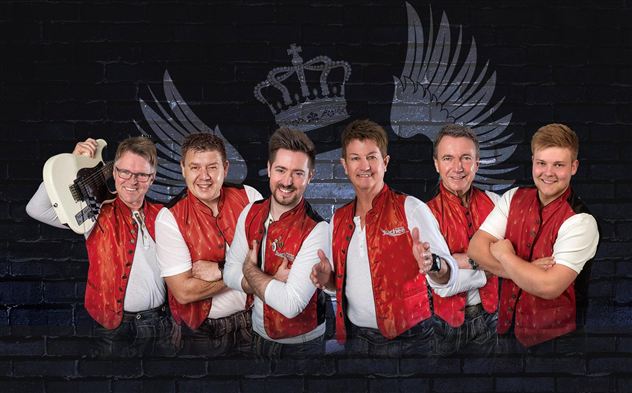 Eine Gruppe von sechs Musikern steht lächelnd vor einer schwarzen Wand mit einem Krone- und Flügelmotiv. Sie tragen rote Westen und zeigen eine freundliche, einladende Haltung.