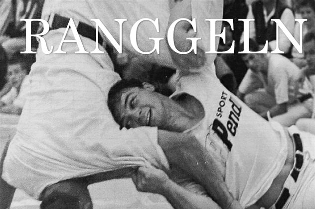 Zwei Männer ringen in einem Wettkampf. Der Sportler auf der rechten Seite lächelt, während er seinen Gegner engagiert hält.