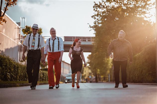 Vier Menschen gehen gemeinsam auf einer Straße, umgeben von Bäumen und Gebäuden. Die Sonne scheint und schafft eine warme Atmosphäre.