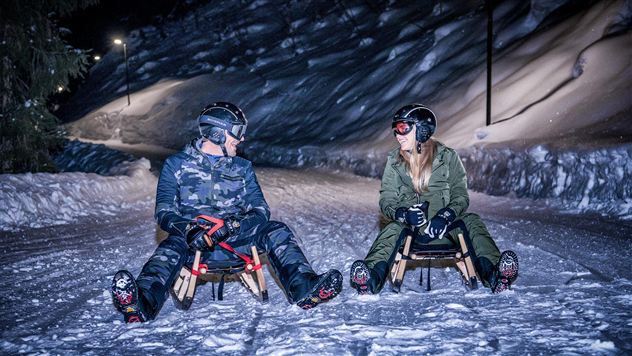 Twee personen zitten op sleetjes in de sneeuw bij nacht. De omgeving is verlicht en een winterse sfeer is zichtbaar.