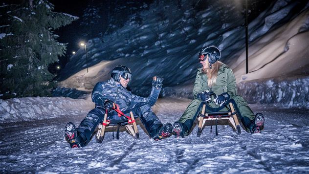 Zwei Personen sitzen im Schnee auf Rodeln und lachen sich an. Die Umgebung ist nachts und mit Schnee bedeckt, umgeben von schneebedeckten Bäumen.