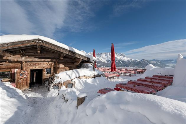 An alpine mountain hut in the snow with red tables outside. The sky is clear and the mountains are visible in the background.