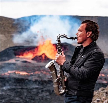 Ein Mann spielt Saxophon vor einem beeindruckenden Vulkan mit sprühender Lava. Die Szenerie ist dramatisch und einzigartig.