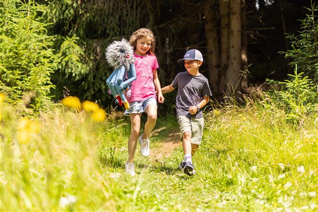 Two children are happily running on a woodland path, surrounded by green plants and flowers. The girl is carrying a toy and smiling as she plays with the boy.
