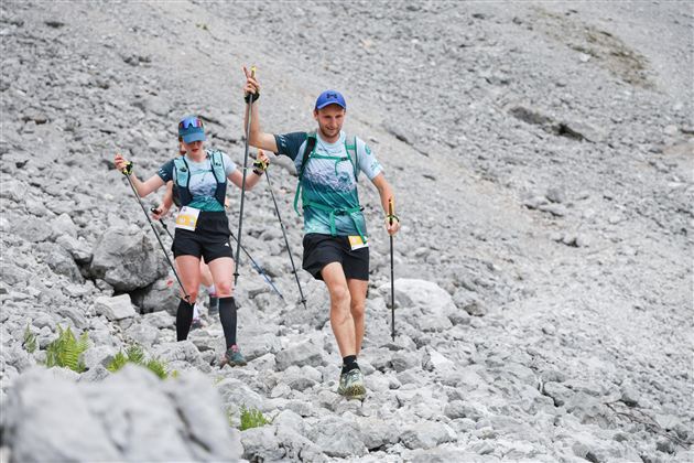 Two athletes are walking with hiking poles over rocky terrain. They are wearing light sports clothing and appear motivated.