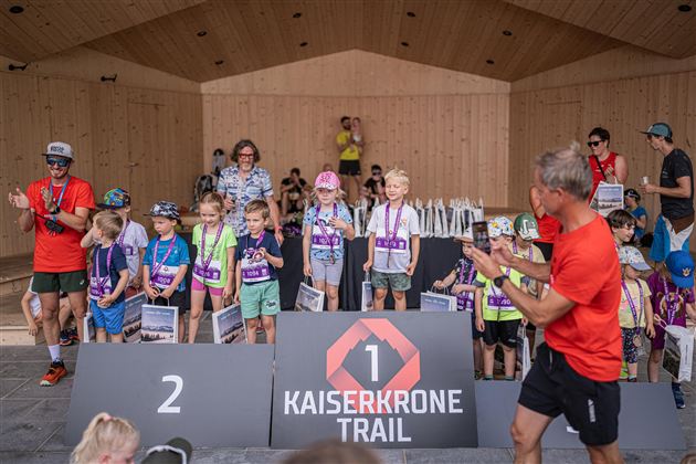 A group of children stands on a stage at the Kaiserkrone Trail. They hold medals and trophies while adults cheer them on.