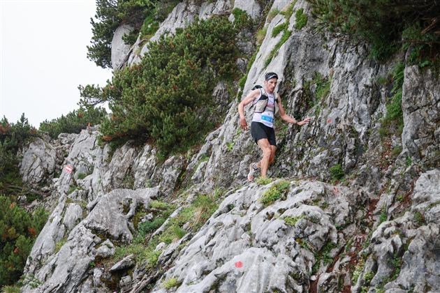 A man is climbing on a rocky path in the mountains. The surroundings are forested and cloudy.
