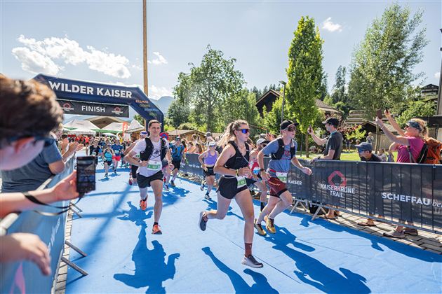 A group of runners reaches the finish line in a race.  
Spectators stand along the course and cheer on the participants.