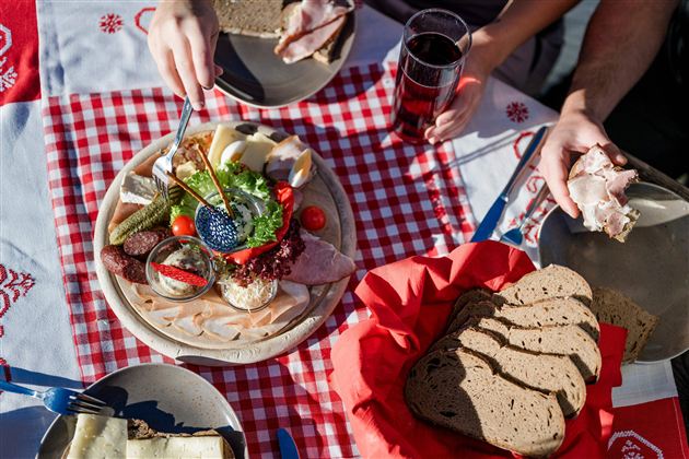 Een kleurrijke plaat met verschillende vlees- en kaaswaren, verse groenten en brood. Op de achtergrond zijn drankjes en andere gerechten op een geruite tafel te zien.