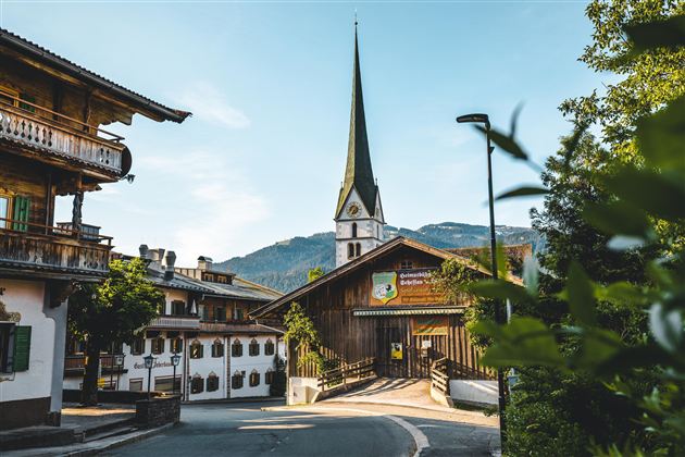 Een schilderachtige dorpsstraat met traditionele architectuur en een hoge kerktoren. Op de achtergrond zijn zachte bergen en een heldere lucht te zien.
