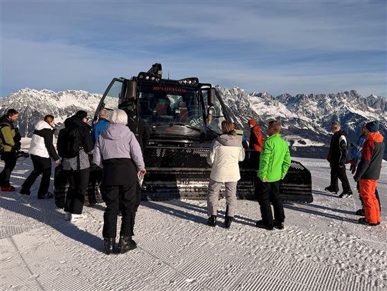 Eine Gruppe von Menschen steht um einen Pistenbully in den Bergen. Im Hintergrund sind schneebedeckte Gipfel und ein klarer Himmel zu sehen.